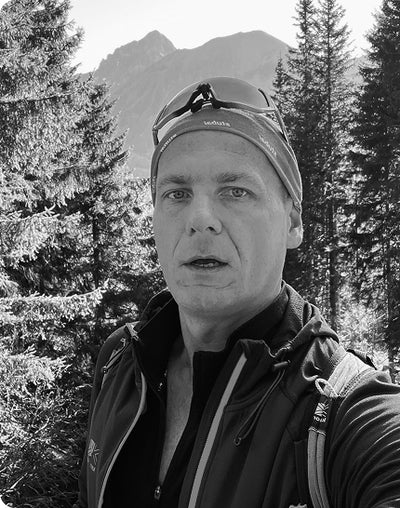 Kyrylo wearing a cap with sunglasses on top, standing in a forest with mountains in the background.